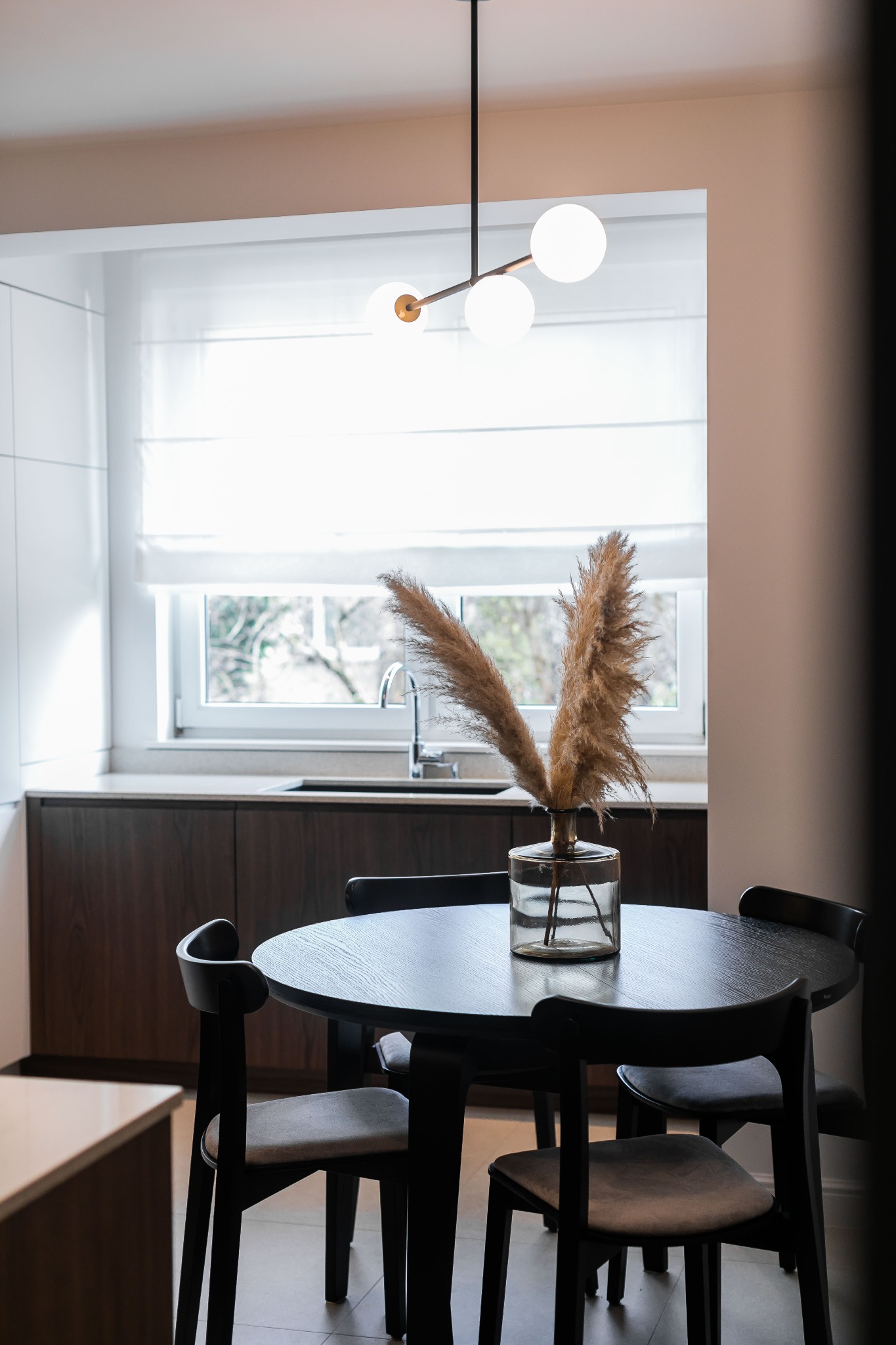 Stół z ciemnego drewna i cztery krzesła w jasnej kuchni z oknem, na stole wazon z trawą pampasową, nad stołem nowoczesna lampa z trzema kulistymi kloszami.