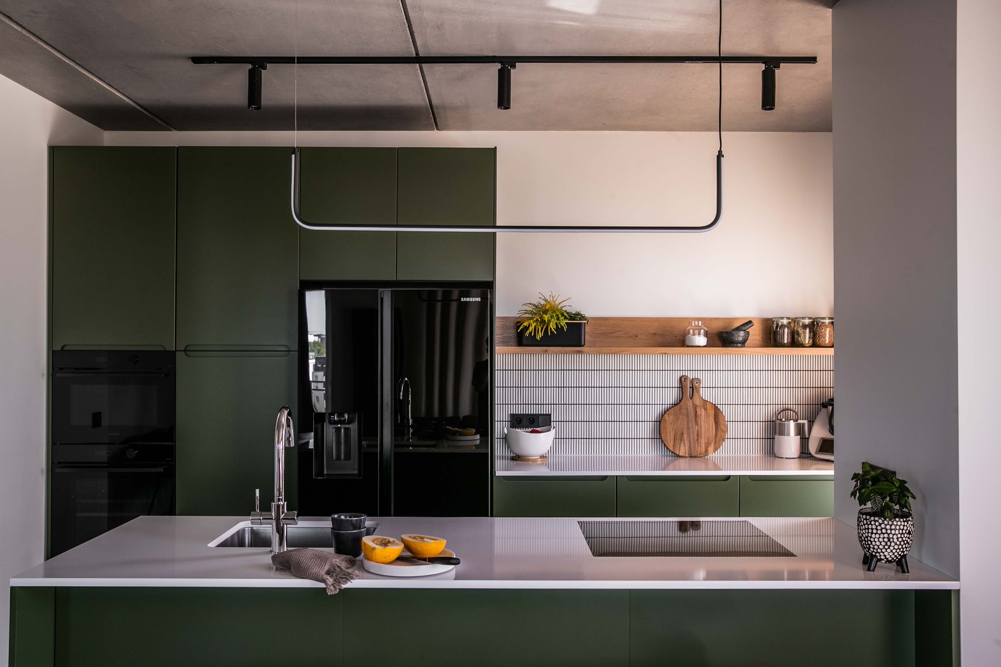 Nowoczesna kuchnia z ciemnozielonymi szafkami, czarną lodówką Samsung, szarą wyspą z owocami, oświetleniem LED i betonowym sufitem.