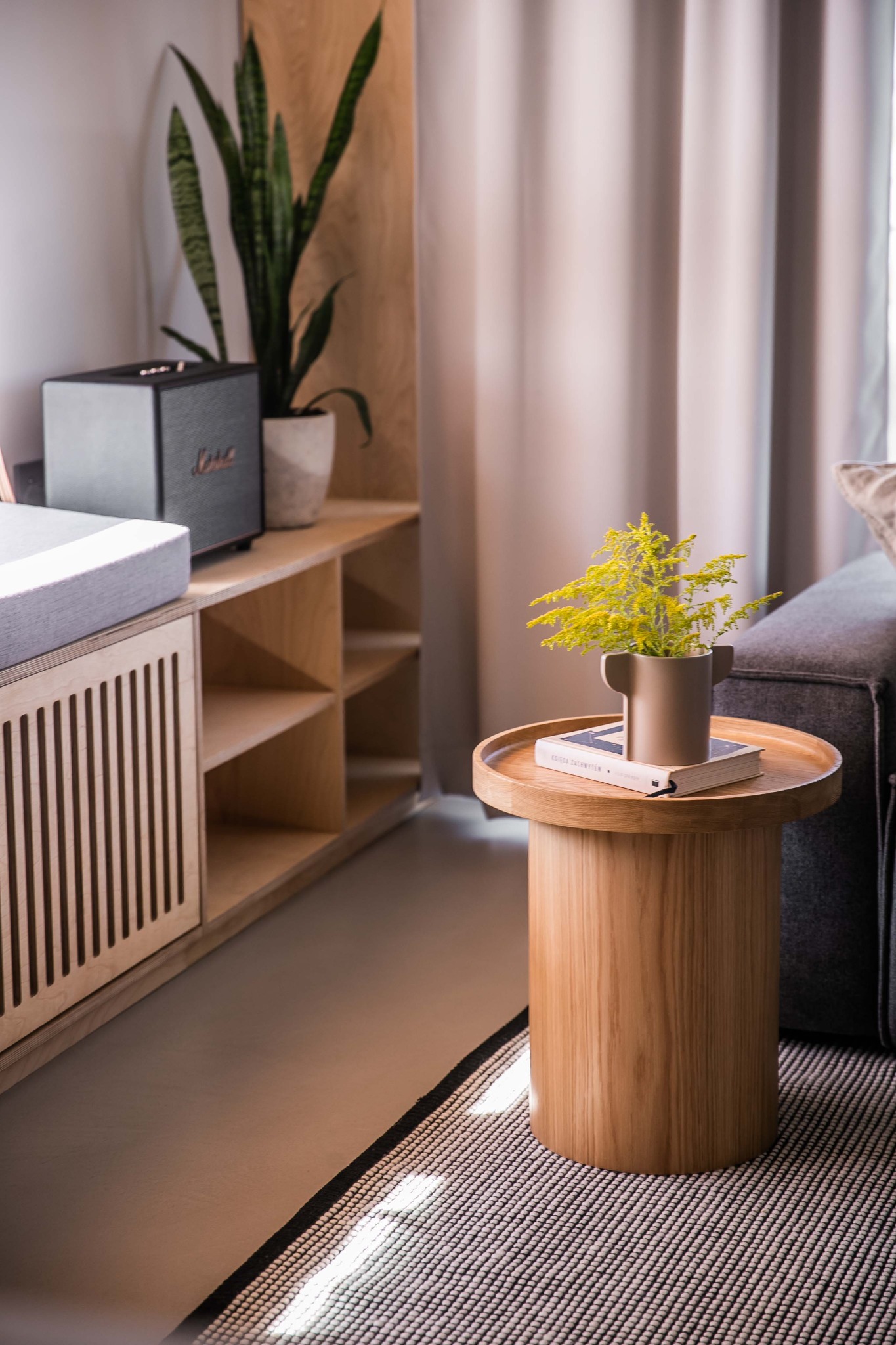 Minimalistyczny salon: drewniany stolik z żółtymi kwiatami i książką, obok szara sofa i dywan w geometryczny wzór, w tle drewniana szafka z głośnikiem Marshall i rośliną w doniczce, całość...