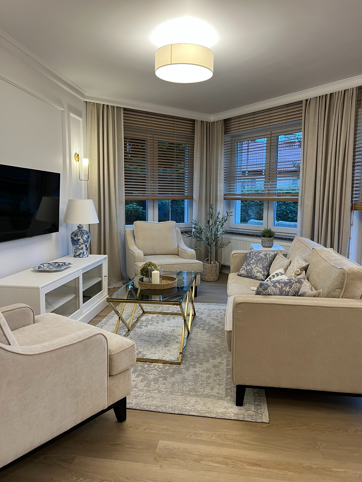 Elegancki salon z beżowymi sofami, szklanym stolikiem kawowym ze złotymi nogami i drewnianymi żaluzjami. Lampy ścienne i dekoracje dopełniają aranżację.