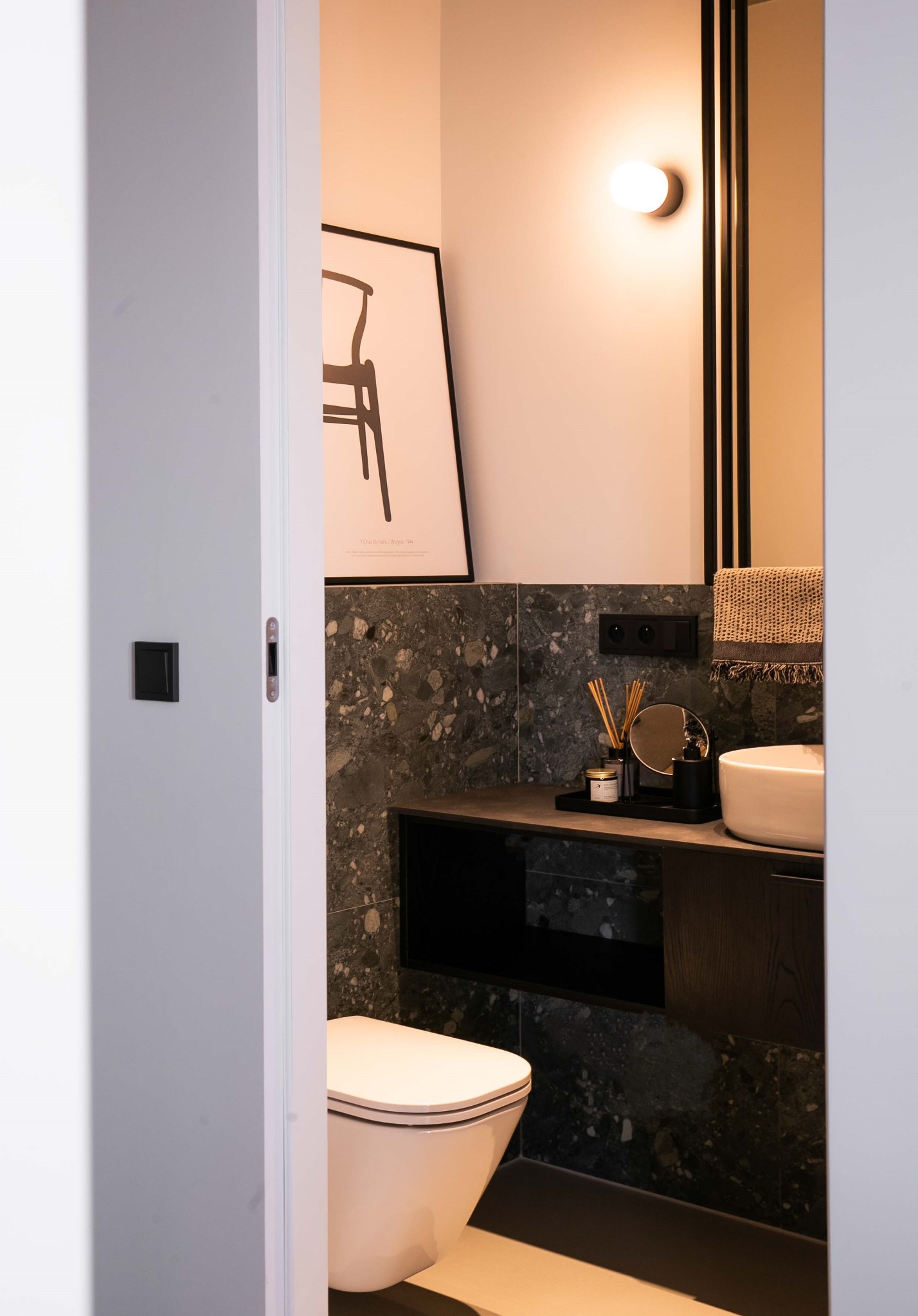 Nowoczesna, mała łazienka z umywalką nablatową, toaletą i dekoracyjną grafiką. Ciemne płytki imitujące kamień i minimalistyczne oświetlenie.