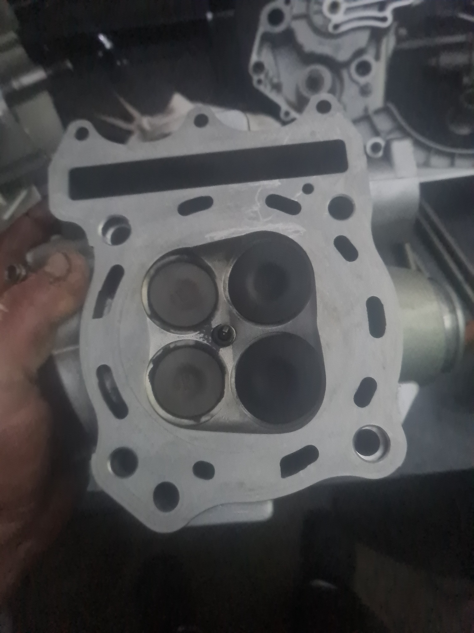Głowica silnika motocyklowego z czterema zaworami, widoczne ślady użytkowania i nagaru, trzymana w dłoni, na drugim planie inne elementy silnika.