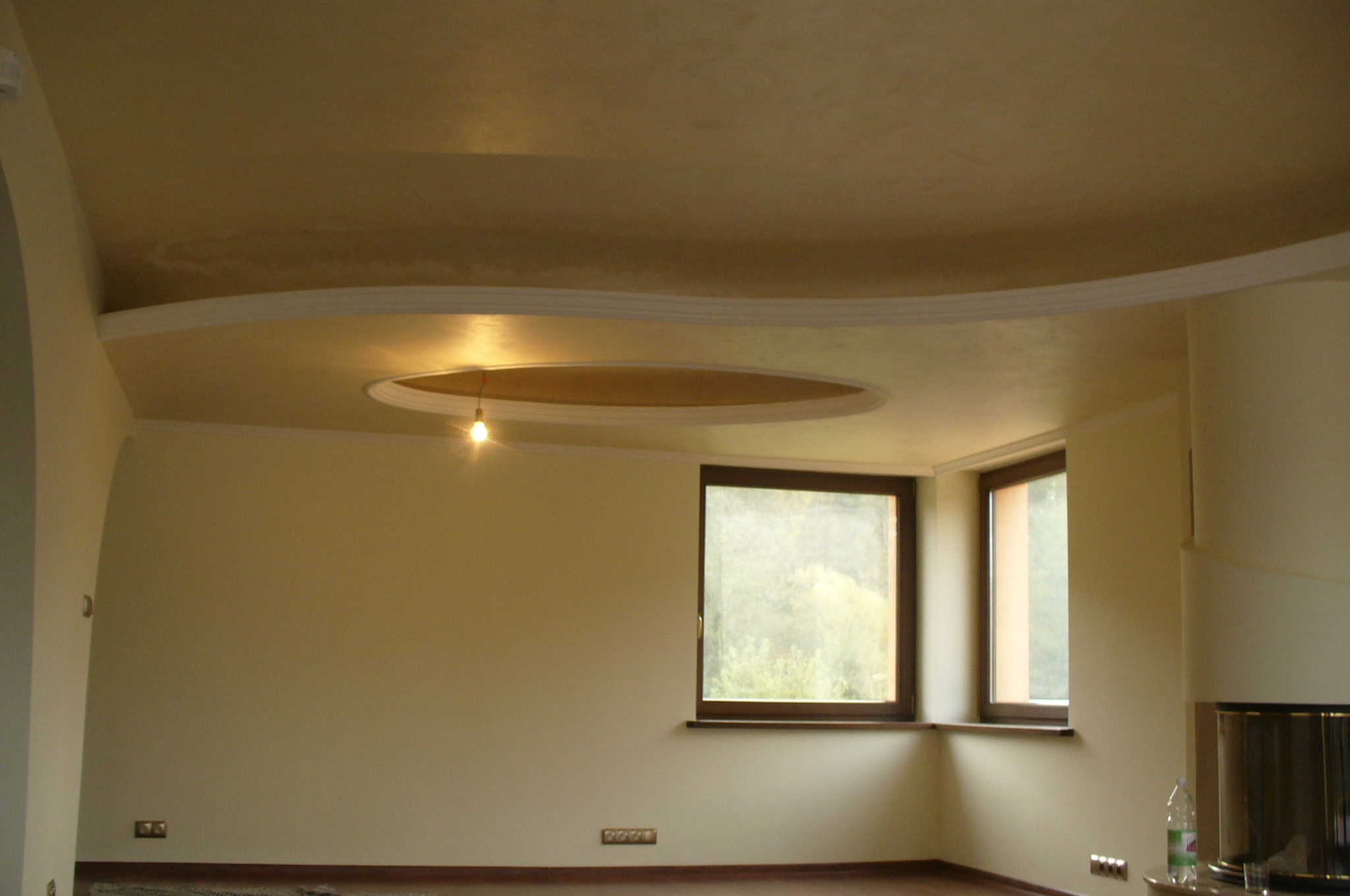 Wnętrze pokoju z kremowymi ścianami, sztukaterią na suficie i owalnym zagłębieniem z żarówką pośrodku, dwa okna narożne z brązowymi ramami, widoczny fragment kominka i podłogi.