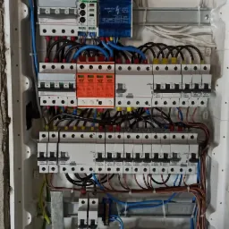 Rozdzielnia elektryczna w białej obudowie, widoczne zabezpieczenia różnicowoprądowe, ograniczniki przepięć, przewody w kolorach niebieskim, żółto-zielonym i brązowym, obudowa wmurowana w surową...