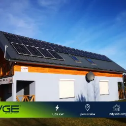Dom z szarą elewacją i panelami słonecznymi na dachu, widoczne okna dachowe, antena satelitarna, rolety zewnętrzne i logo firmy PVGE na dole zdjęcia.