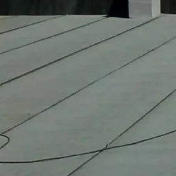 Szary, płaski dach z papy termozgrzewalnej z widocznym kominem wentylacyjnym i anteną satelitarną w tle, zarys zabudowań oraz zieleń drzew.