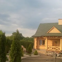 Nowo wybudowany, drewniany dom z zielonym dachem z dachówki w Poznaniu, otoczony tujami i kamieniami dekoracyjnymi, z widoczną taczką i stertą materiałów budowlanych.