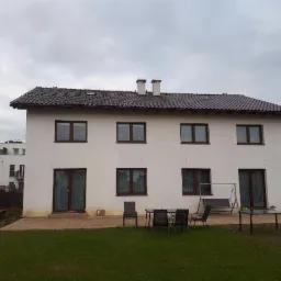 Dwupiętrowy dom z brązowym dachem z dachówki ceramicznej, widok z ogrodu w pochmurny dzień. Widoczne kominy i okna z brązowymi ramami.