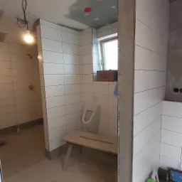 Wnętrze łazienki w trakcie remontu, widoczne białe płytki na ścianach, surowy sufit, zamontowany pisuar i prowizoryczny stołek, narzędzia w skrzynce w rogu, w tle kabina prysznicowa.