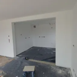 Wnętrze pokoju po świeżym pomalowaniu ścian na biało, widoczne przygotowania do dalszych prac wykończeniowych, zabezpieczona podłoga, wystające kable i puszki elektryczne.