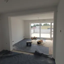 Wnętrze pokoju po świeżym pomalowaniu ścian na biało, z widocznymi puszkami farby, wiaderkami i zabezpieczeniem podłogi folią malarską, otwarte okno z widokiem na zabudowę mieszkalną i balkon...