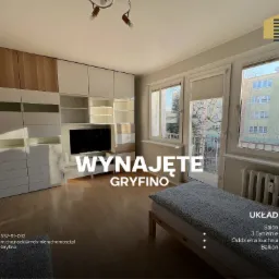 Jasne mieszkanie z balkonem, widok na miasto przez okna, zabudowa meblowa z telewizorem, informacja o wynajmie nałożona cyfrowo na zdjęcie.