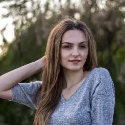 Portret młodej kobiety z długimi brązowymi włosami, ubrana w szary sweter, pozująca na tle rozmytych drzew z delikatnym oświetleniem.