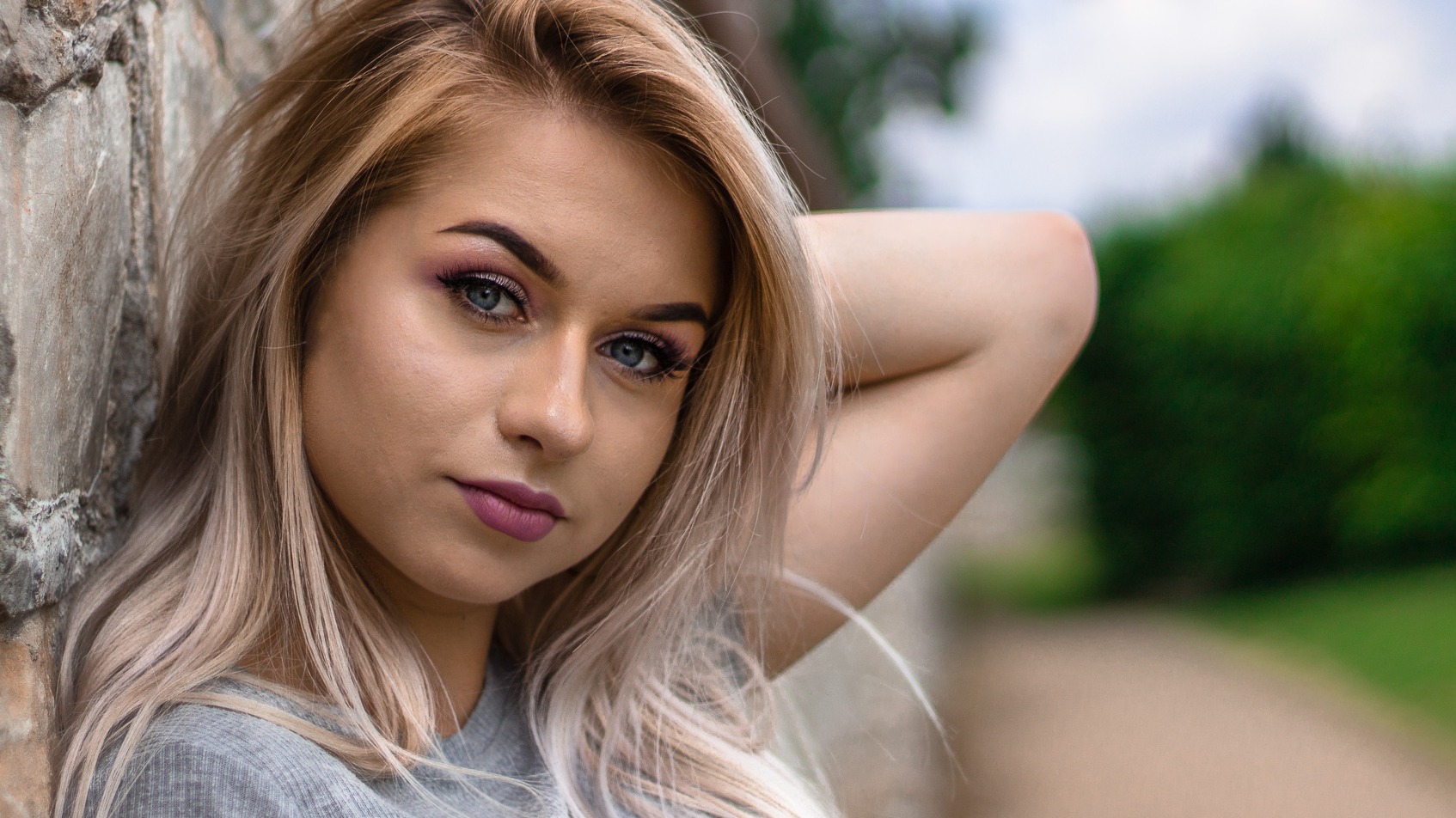 Portret młodej kobiety z blond włosami opierającej się o kamienną ścianę, z delikatnym makijażem w odcieniach różu i fioletu, z zamyślonym spojrzeniem skierowanym w obiektyw.