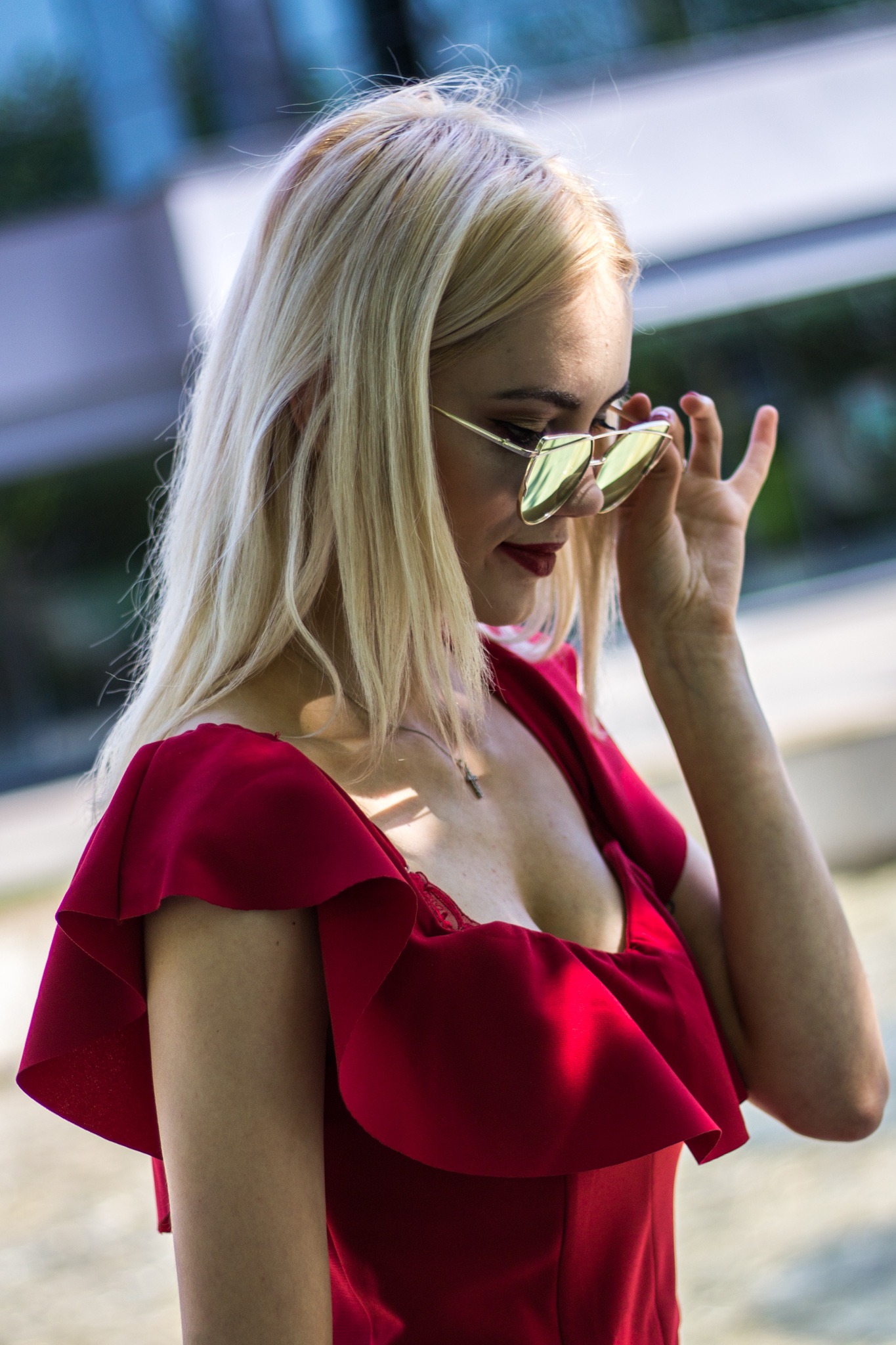 Portret kobiety z blond włosami w czerwonej sukience z falbanami, poprawiającej okulary przeciwsłoneczne o złotych oprawkach.