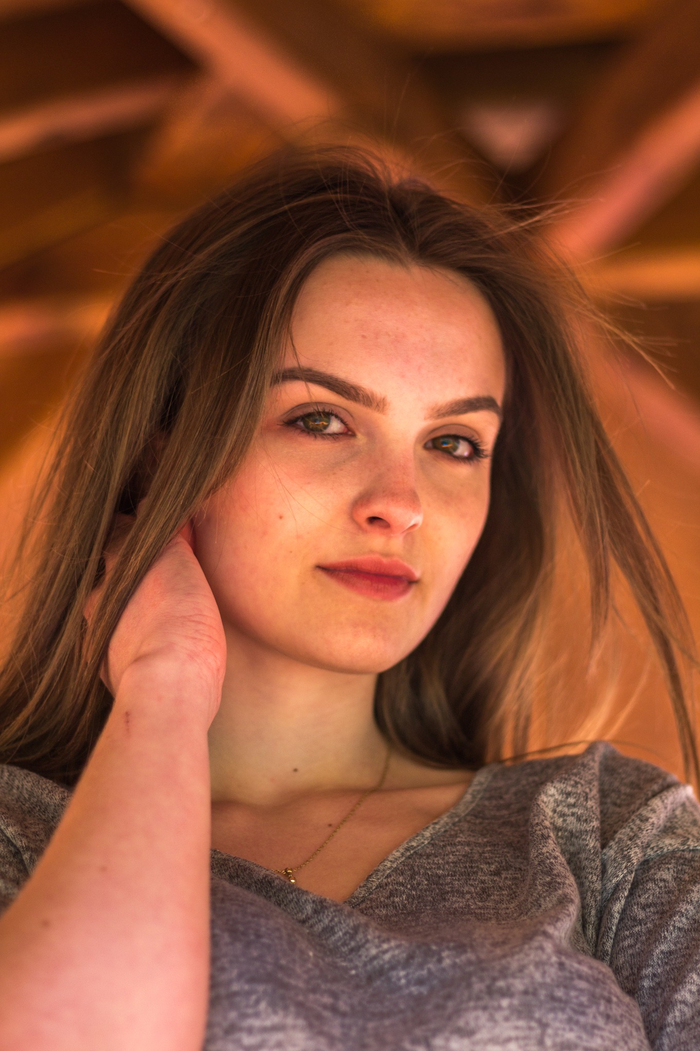 Portret młodej kobiety z delikatnym makijażem, w szarej bluzce, z dłonią przy twarzy, sfotografowanej w Piasecznie z naturalnym oświetleniem.