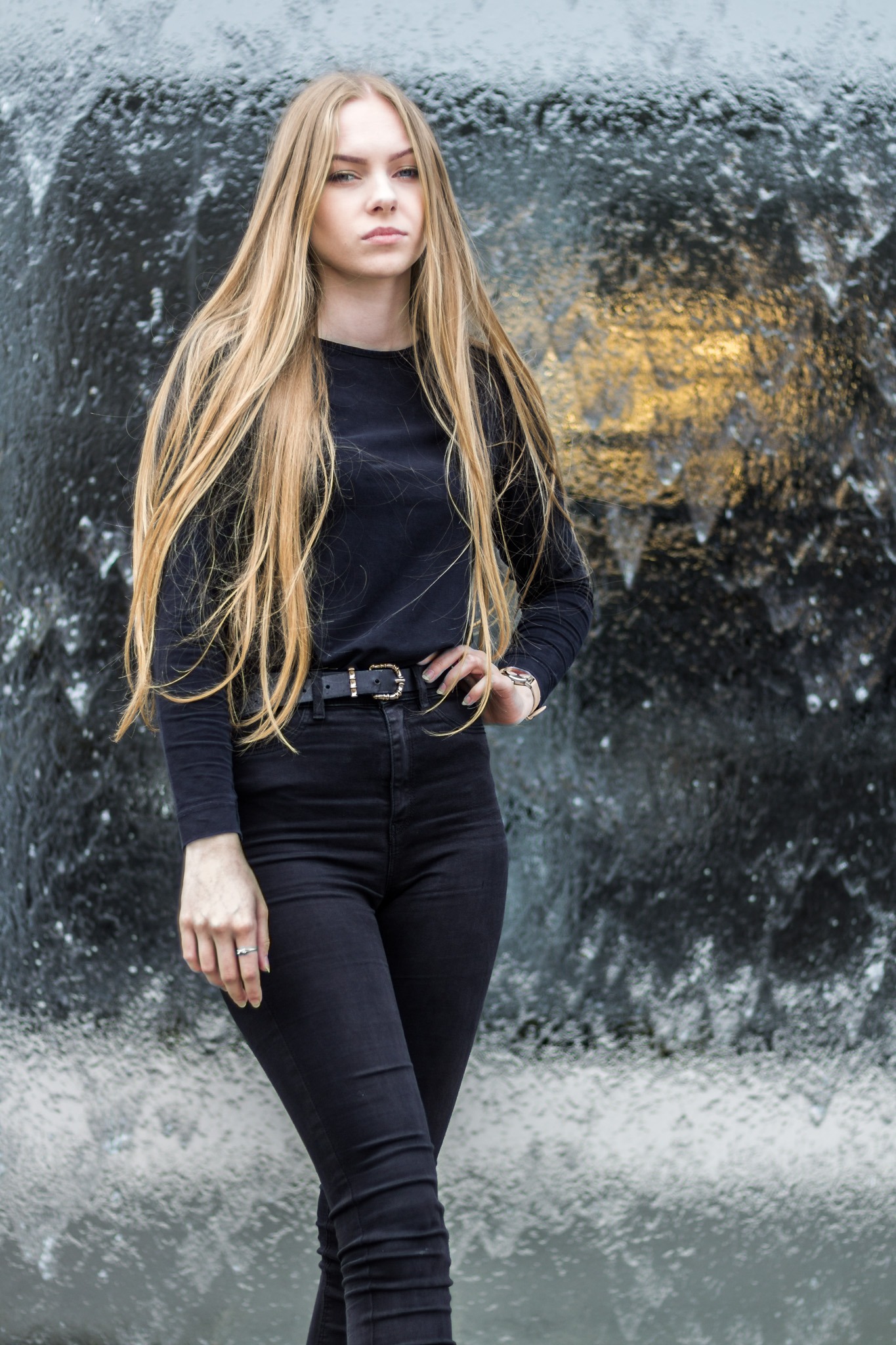 Portret kobiety z długimi blond włosami, ubrana w czarny strój, pozująca na tle wodnej ściany z delikatnym oświetleniem.