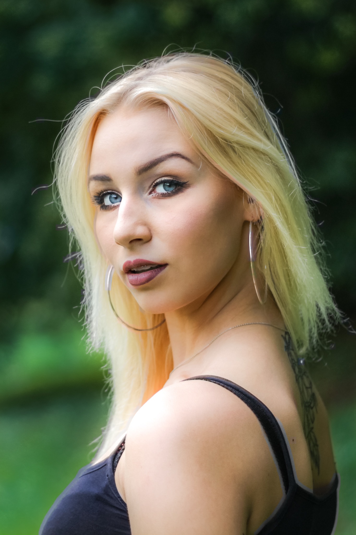 Portret kobiety z blond włosami, niebieskimi oczami i tatuażem na plecach, spoglądającej przez ramię, z dużymi srebrnymi kolczykami kołami