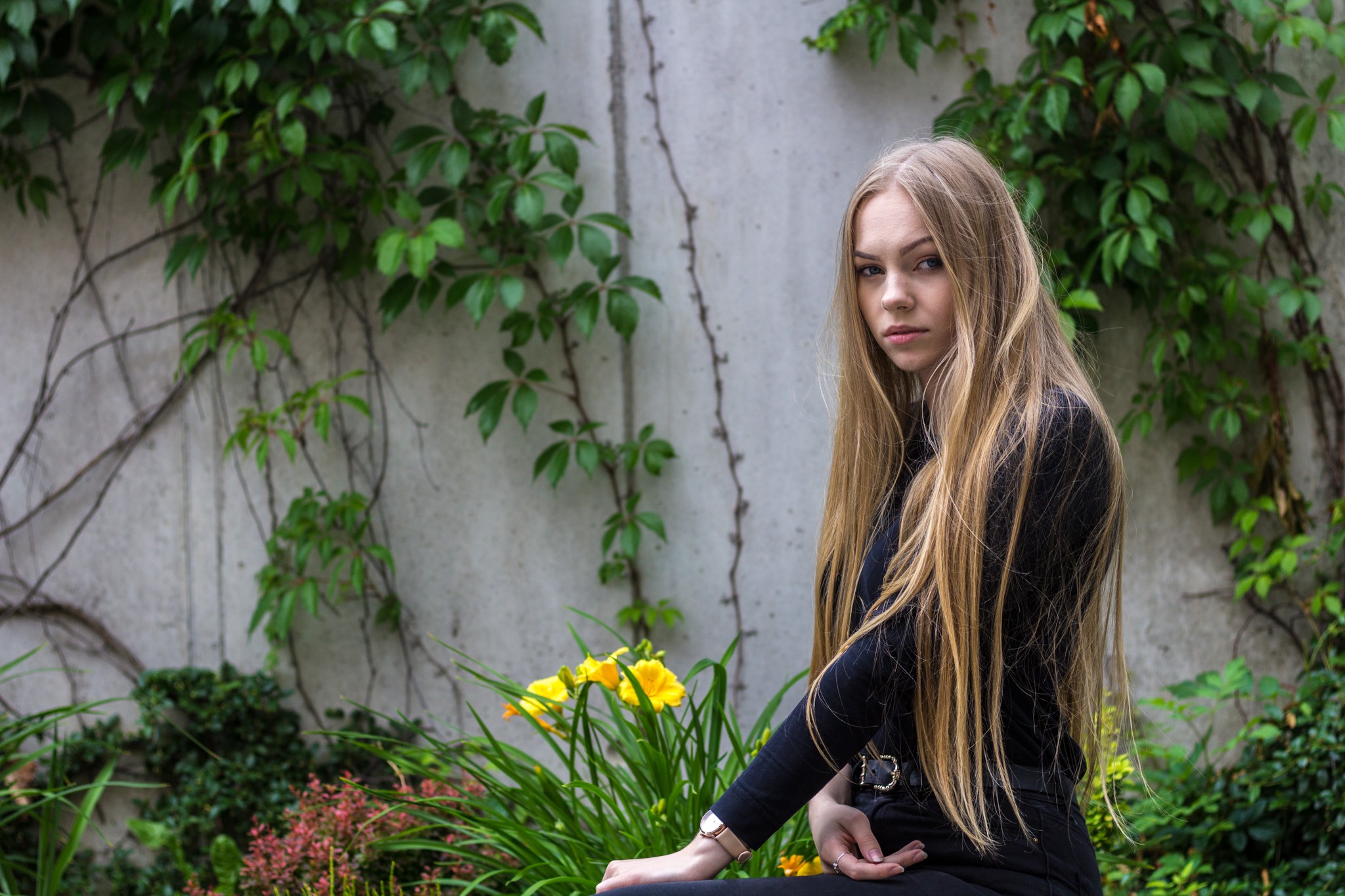 Portret młodej kobiety z długimi blond włosami, ubrana na czarno, siedzącej na tle betonowej ściany porośniętej bluszczem i kwitnącymi żółtymi liliowcami.