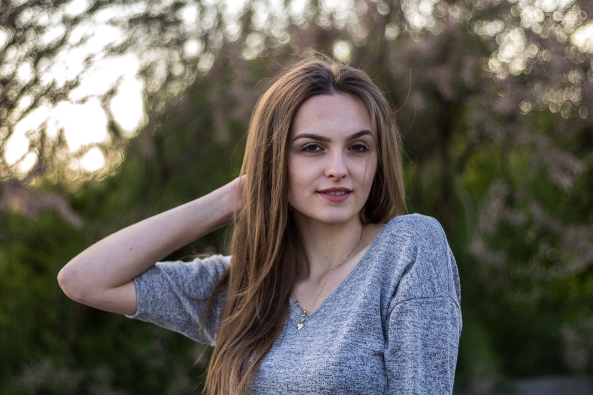 Portret młodej kobiety z długimi brązowymi włosami, ubrana w szary sweter, pozująca na tle rozmytych drzew z delikatnym oświetleniem.