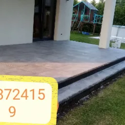 Wykończony taras z szarych płyt z dwoma stopniami, widok na trawnik, basen i domek dla dzieci w tle.