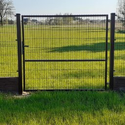PHU FELIX Weronika Kazimierska - Nowoczesna brama z metalowej siatki w kolorze antracytowym, z widocznym zamkiem i klamką, zamontowana na tle zielonego trawnika i pola, uzupełniona o podmurówkę imitującą kamień.