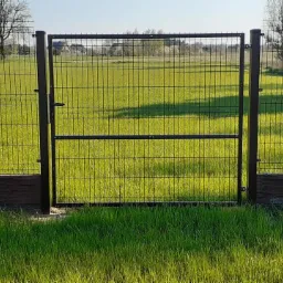 Nowoczesna brama z metalowej siatki w kolorze antracytowym, z widocznym zamkiem i klamką, zamontowana na tle zielonego trawnika i pola, uzupełniona o podmurówkę imitującą kamień.