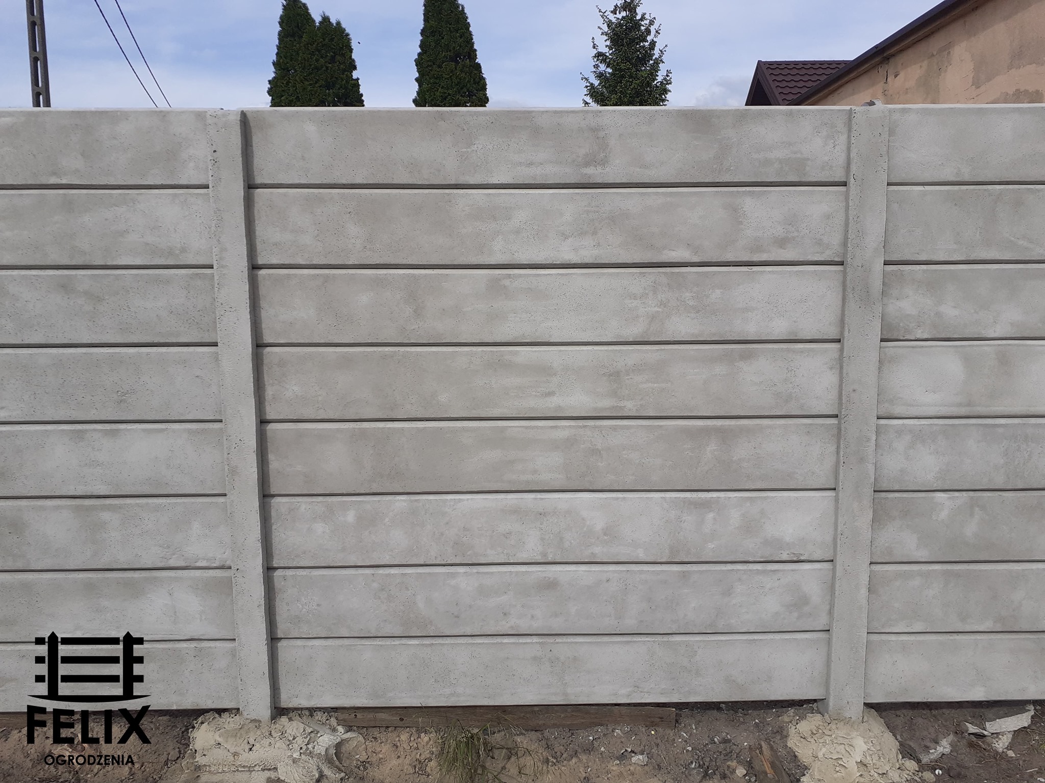 Szary, prefabrykowany płot betonowy z poziomymi panelami, widoczne słupy nośne, w tle drzewa i budynek z brązowym dachem, u dołu logo firmy Felix Ogrodzenia.