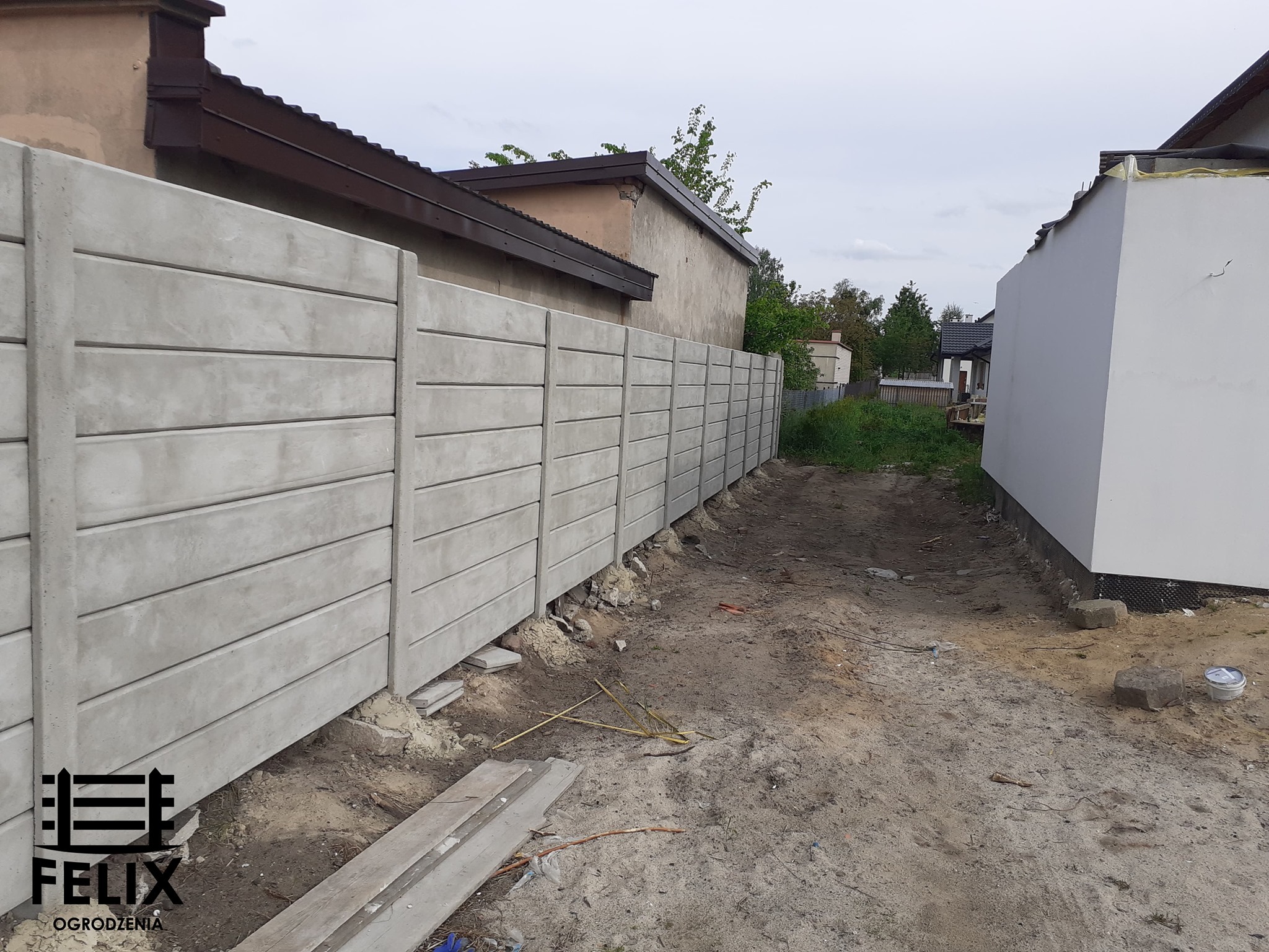 Długi, szary, betonowy płot panelowy z poziomymi przerwami, świeżo zamontowany, z widocznymi fundamentami i logo firmy w lewym dolnym rogu.