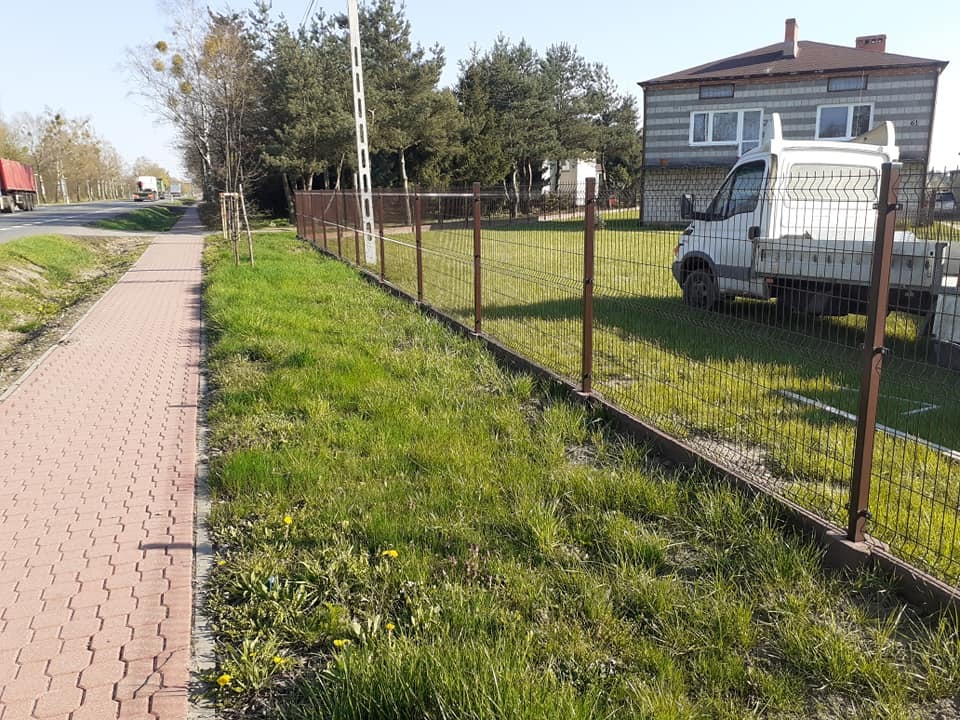 Nowoczesne ogrodzenie z siatki z brązowymi słupkami i podmurówką, oddzielające chodnik z kostki brukowej od trawnika, na tle domu i samochodu dostawczego.