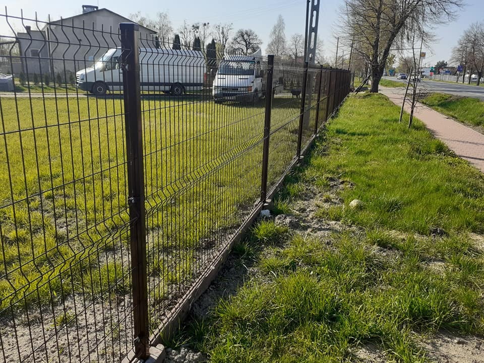 Nowoczesne ogrodzenie z paneli zgrzewanych w kolorze ciemnego brązu, zamontowane na betonowej podmurówce, widok z perspektywy trawnika w kierunku ulicy i zaparkowanych samochodów dostawczych.