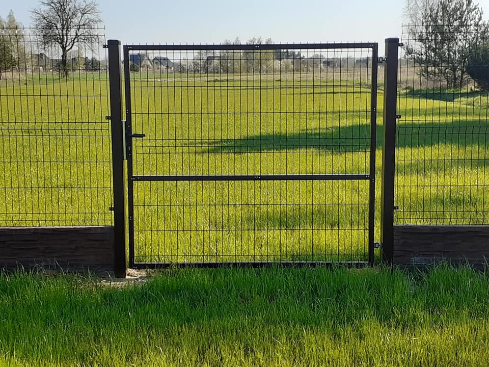 Nowoczesna brama z metalowej siatki w kolorze antracytowym, z widocznym zamkiem i klamką, zamontowana na tle zielonego trawnika i pola, uzupełniona o podmurówkę imitującą kamień.