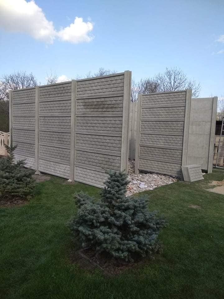 Fragmenty betonowego ogrodzenia z fakturą cegły, ustawione na trawniku przed montażem, z widocznymi młodymi świerkami i elementami gruzu.