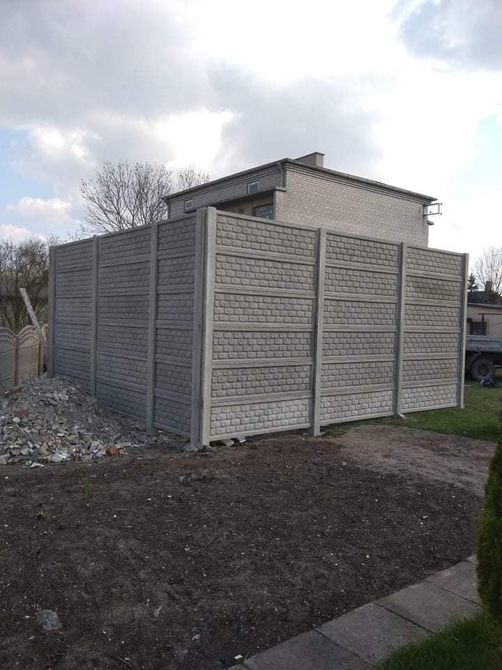Nowo postawione, szare ogrodzenie z prefabrykatów betonowych o wzorze cegieł, widoczne na tle domu i pochmurnego nieba, z niewielkim stosem gruzu u podstawy.