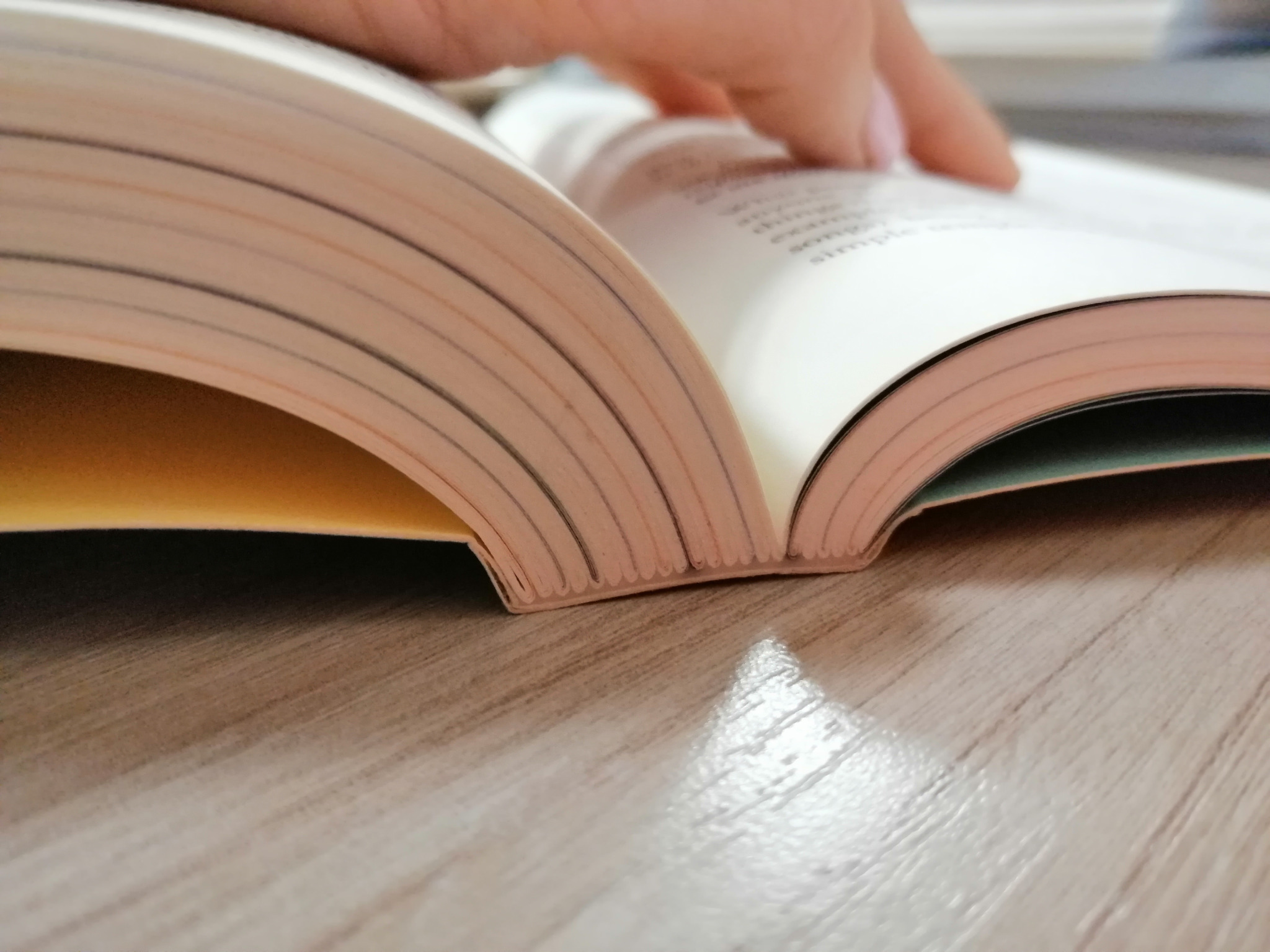 Otwarta książka w miękkiej oprawie szyto-klejonej, widoczne zszycia na grzbiecie, druk cyfrowy, delikatny cień na drewnianym blacie.