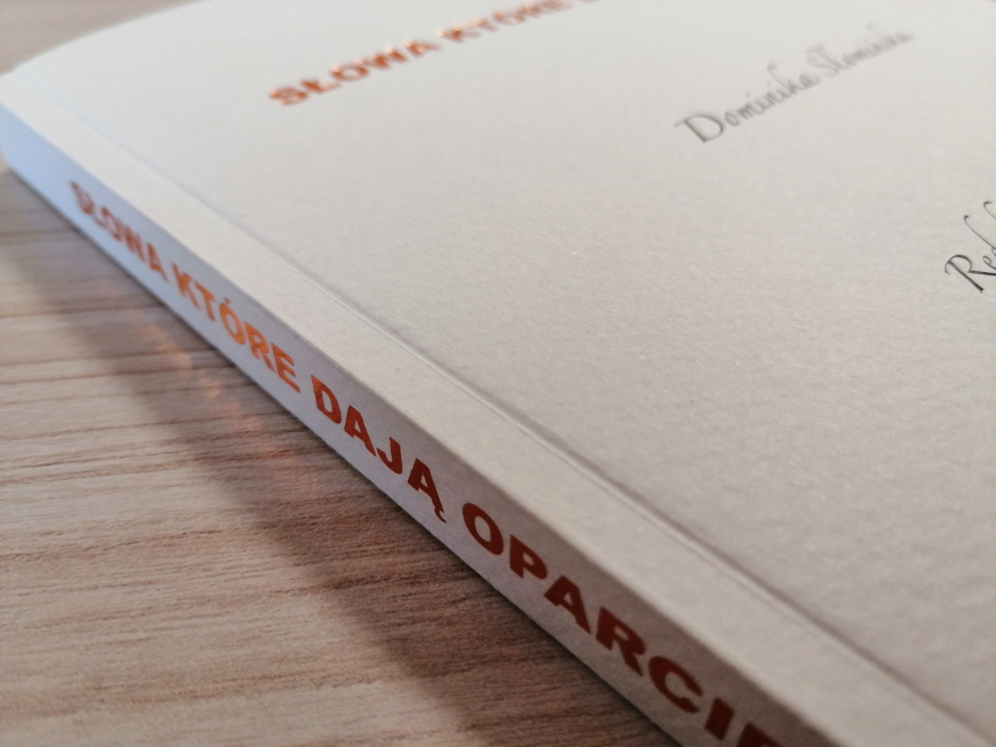 Książka w miękkiej oprawie, z tytułem wytłoczonym na okładce i grzbiecie w kolorze miedzianym, leżąca na drewnianym blacie.