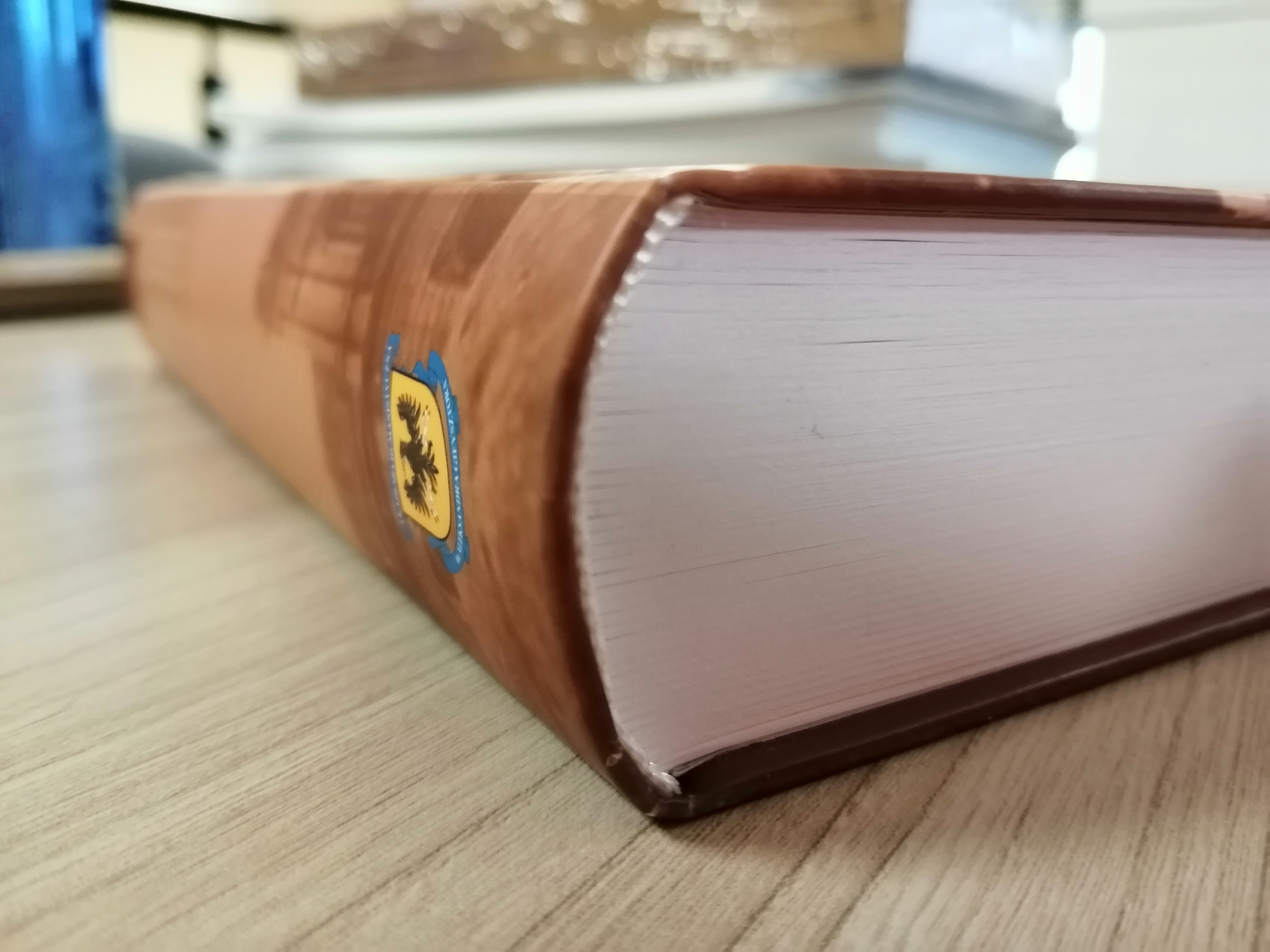 Książka w twardej oprawie z widocznym szyto-klejonym blokiem, leżąca na drewnianym blacie, z herbem na okładce.