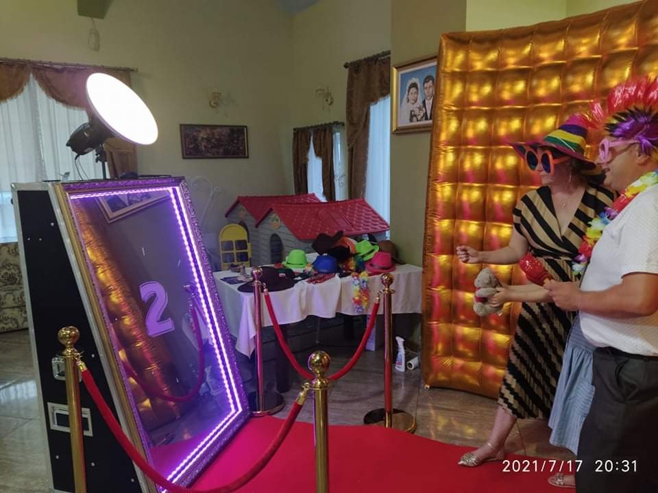 Uczestnicy imprezy pozują do zdjęcia w fotobudce lustrzanej z fioletowym oświetleniem LED, na czerwonym dywanie i ze złotym tłem, rekwizyty na stole w tle.