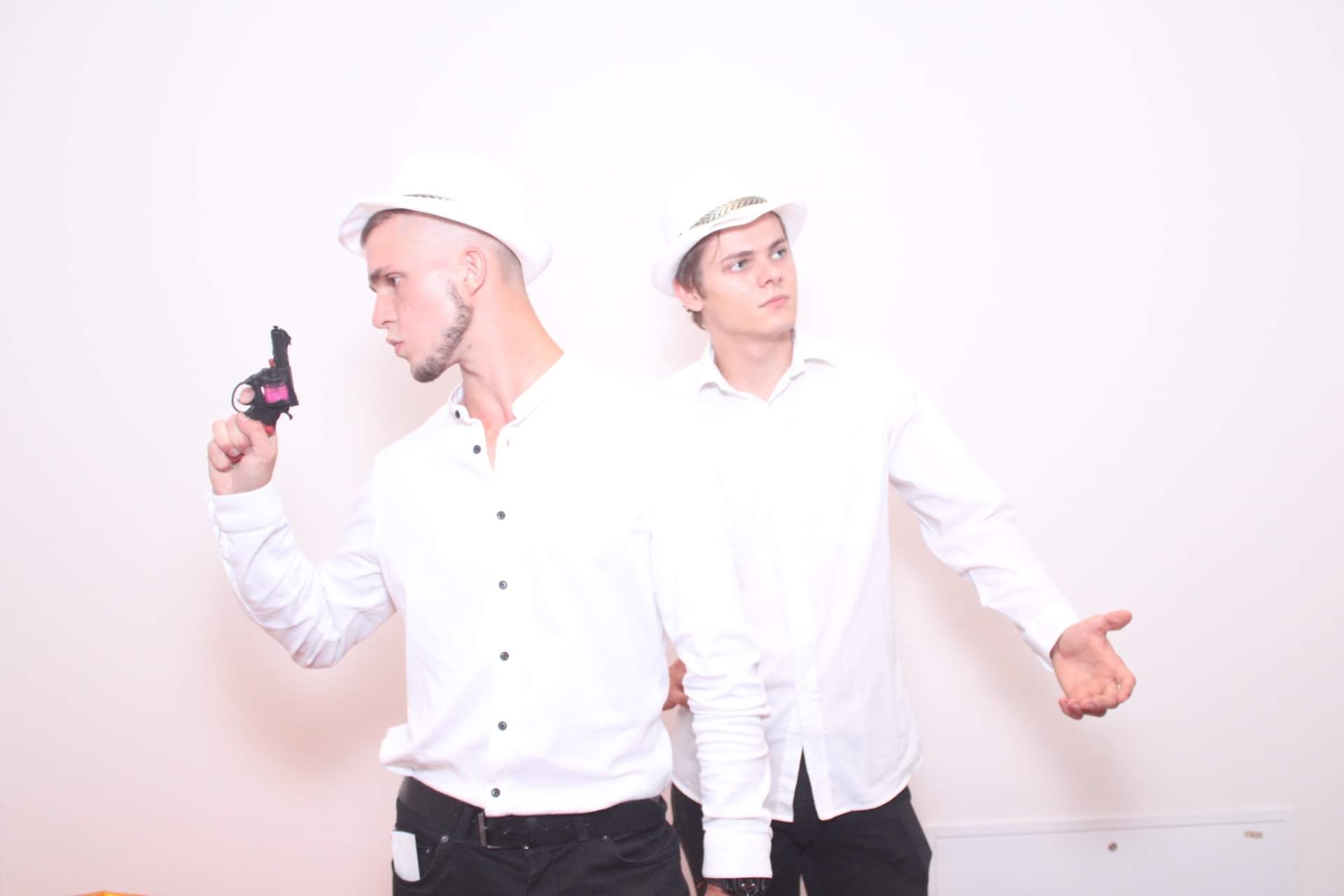 Dwóch mężczyzn w białych koszulach i kapeluszach pozuje z zabawką pistoletem na jasnym tle, sugerując tematyczną sesję zdjęciową lub imprezę.