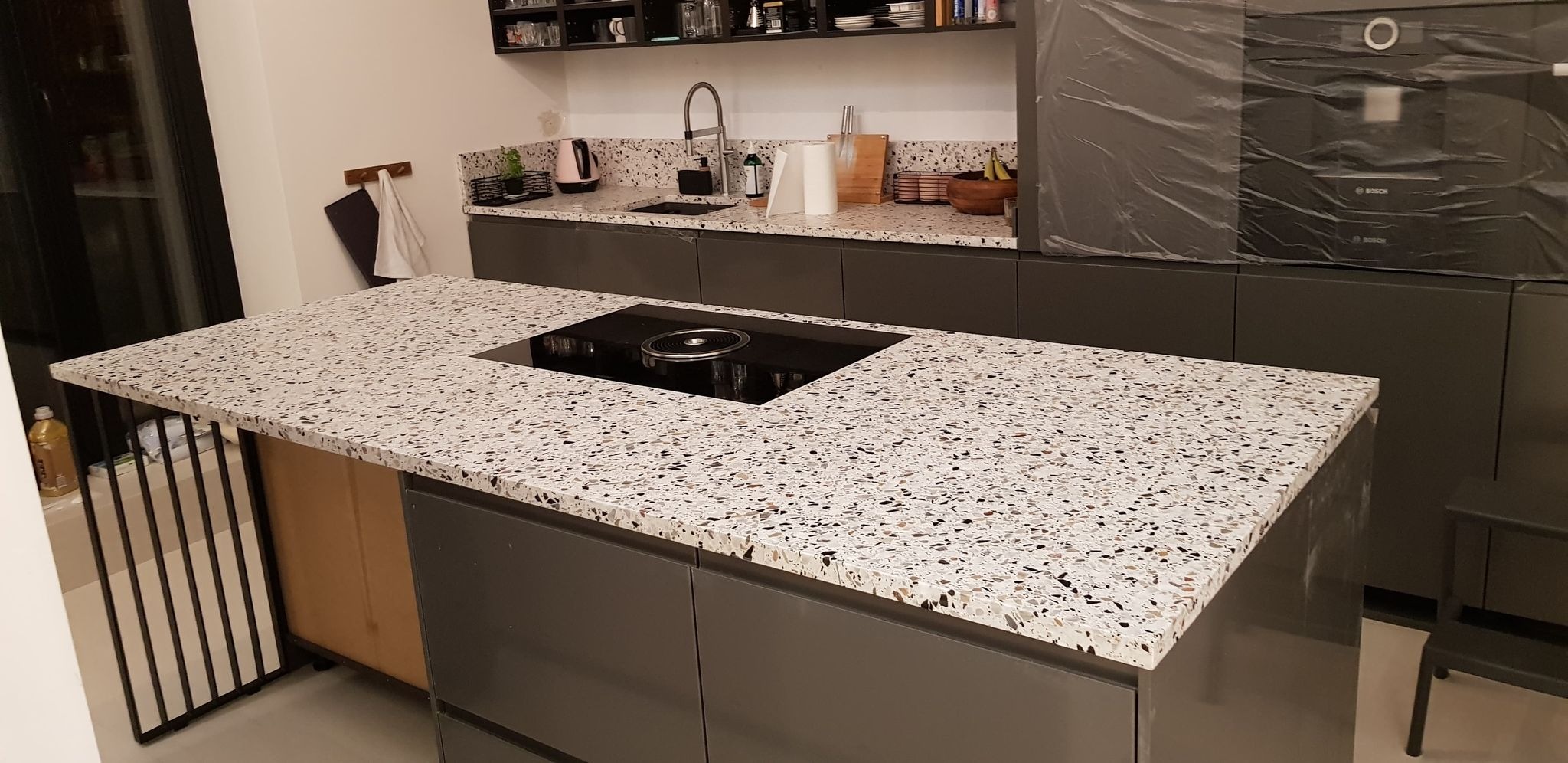 Kuchenny blat w stylu lastryko z wbudowaną płytą indukcyjną, szare szafki kuchenne, w tle zlewozmywak i sprzęt AGD w folii ochronnej.