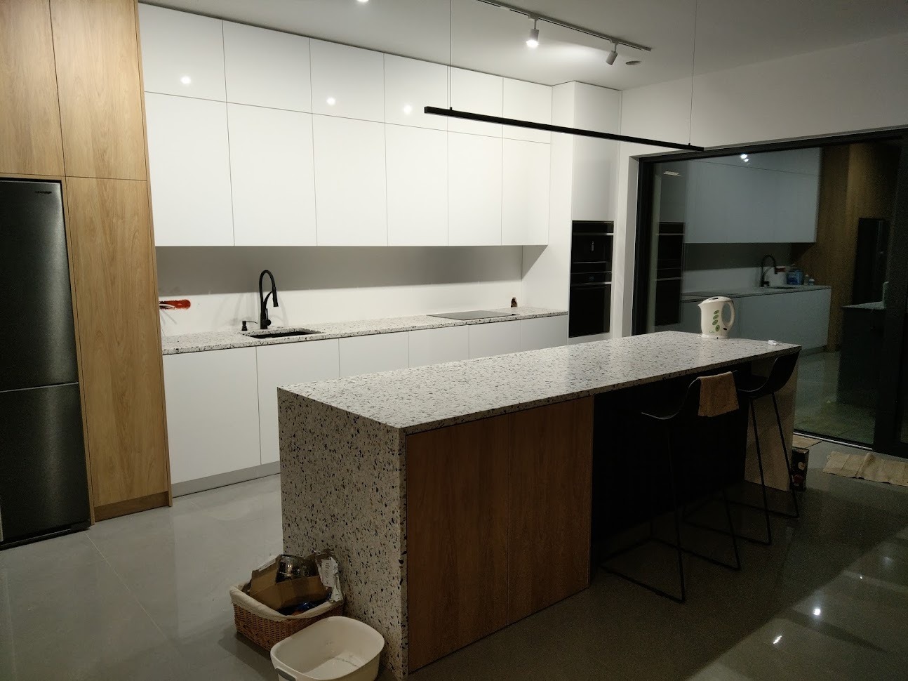 Nowoczesna kuchnia z białymi szafkami i blatem z lastryko, czarna armatura, wyspa kuchenna z drewnianym wykończeniem, widok na drugą kuchnię przez szklane drzwi.