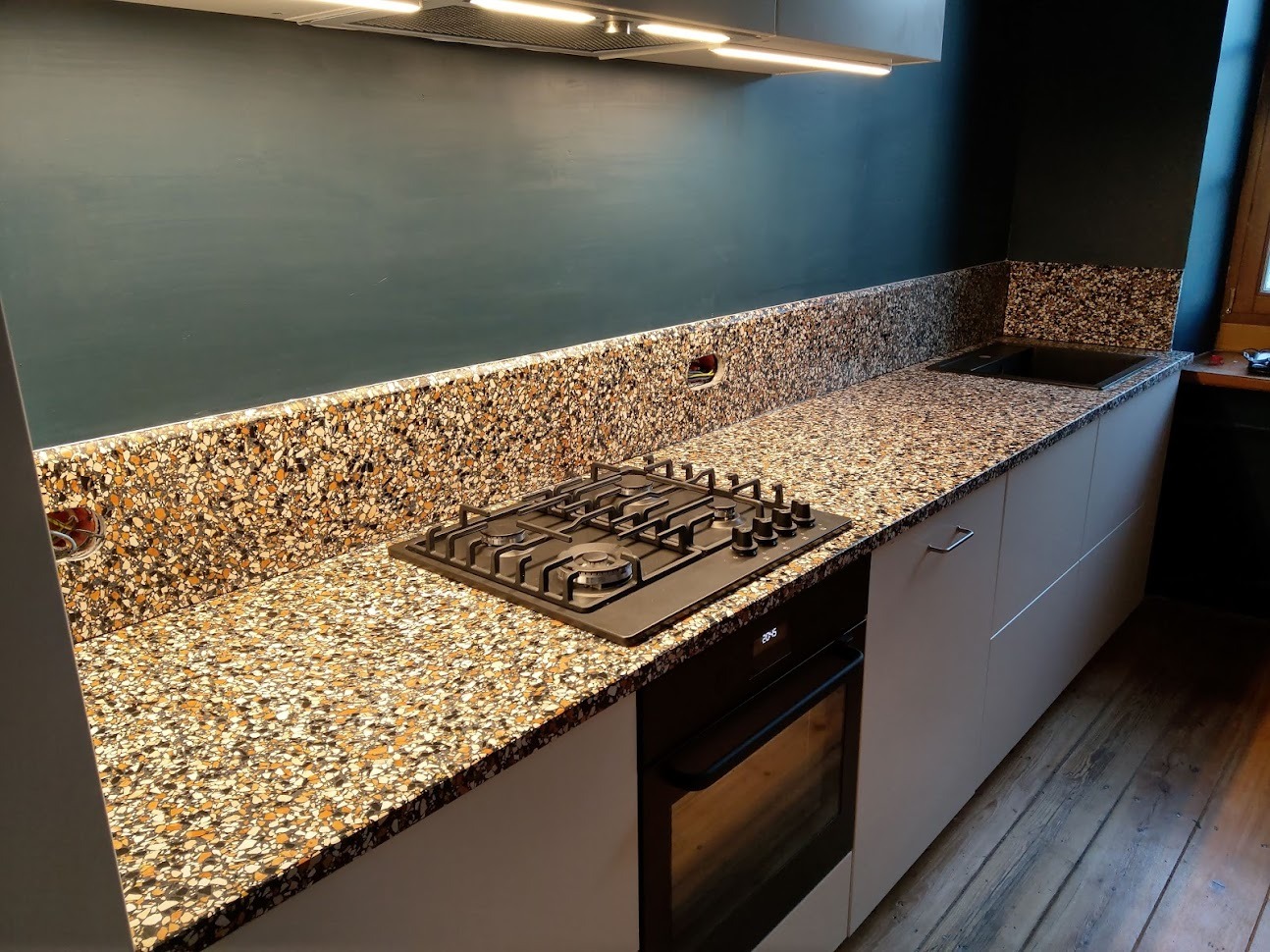 Kuchnia z blatem i okładziną ścienną typu lastryko w odcieniach brązu i beżu, z zamontowaną płytą gazową i piekarnikiem.