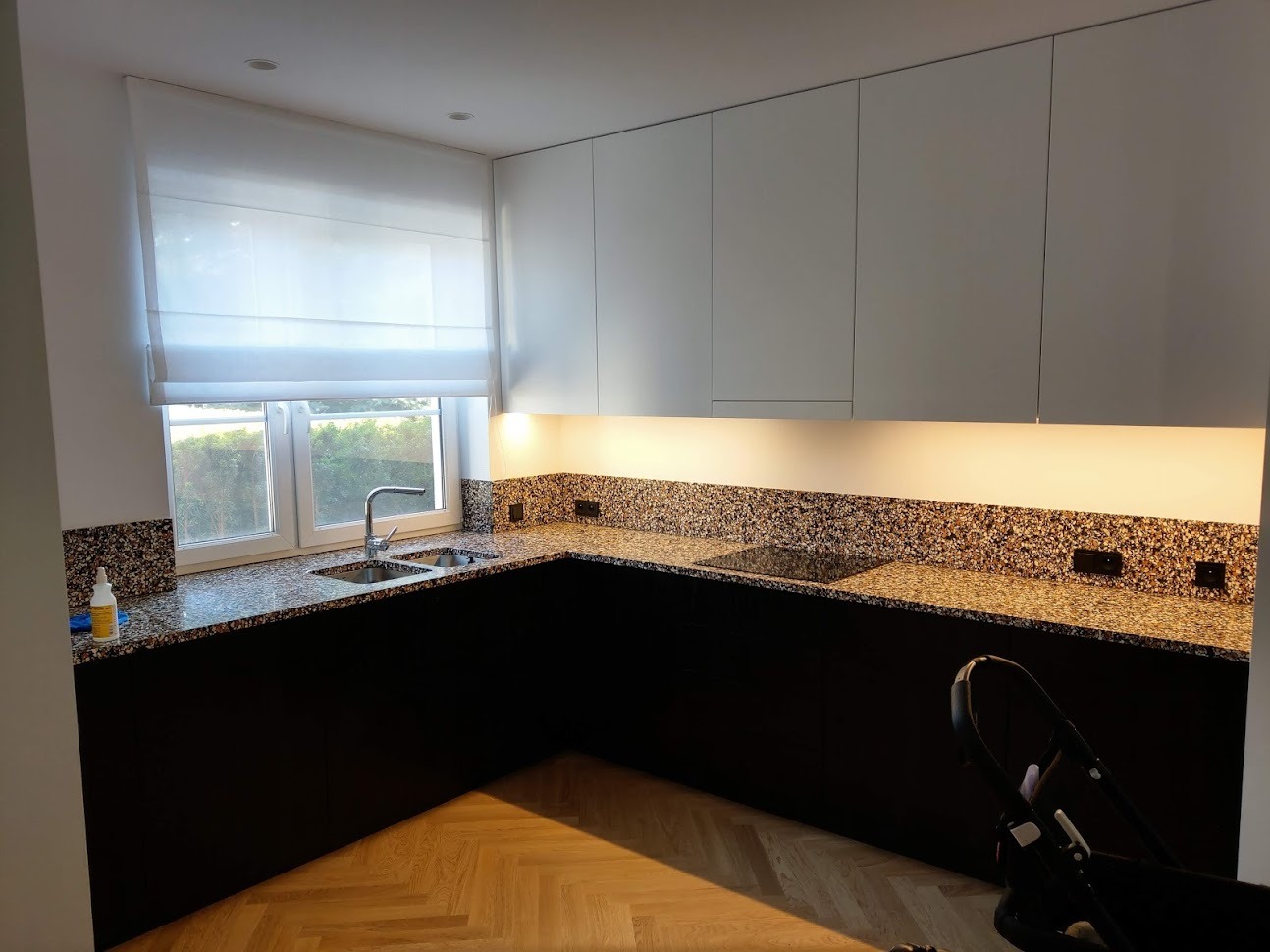 Kuchnia z blatem z lastryko w odcieniach brązu, czarne szafki dolne i białe górne, oświetlenie LED pod szafkami, widok na okno z roletą rzymską.