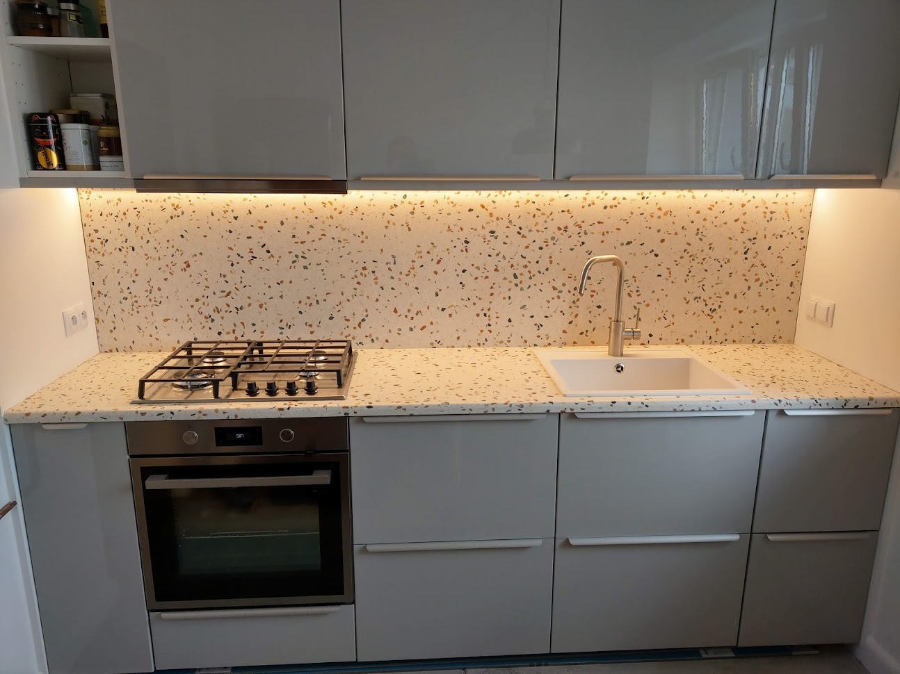 Kuchnia z blatem i ścianą w stylu lastryko, z płytą gazową i zlewozmywakiem, fronty szafek w kolorze szarym.