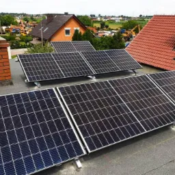 Panele słoneczne zamontowane na płaskim dachu budynku, z widocznymi kominami z cegły i dachami sąsiednich domów w tle.