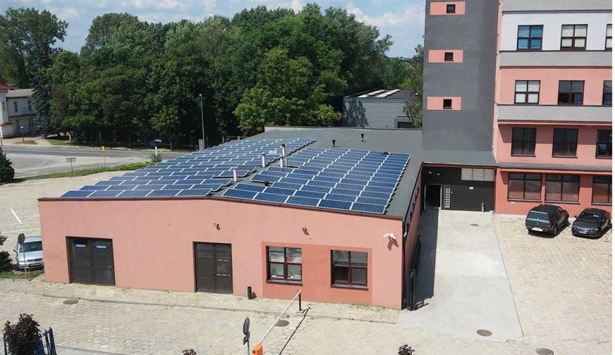 Widok z góry na budynek z zainstalowanymi panelami słonecznymi na dachu, obok budynek wielopiętrowy, drzewa w tle i parking z samochodami.