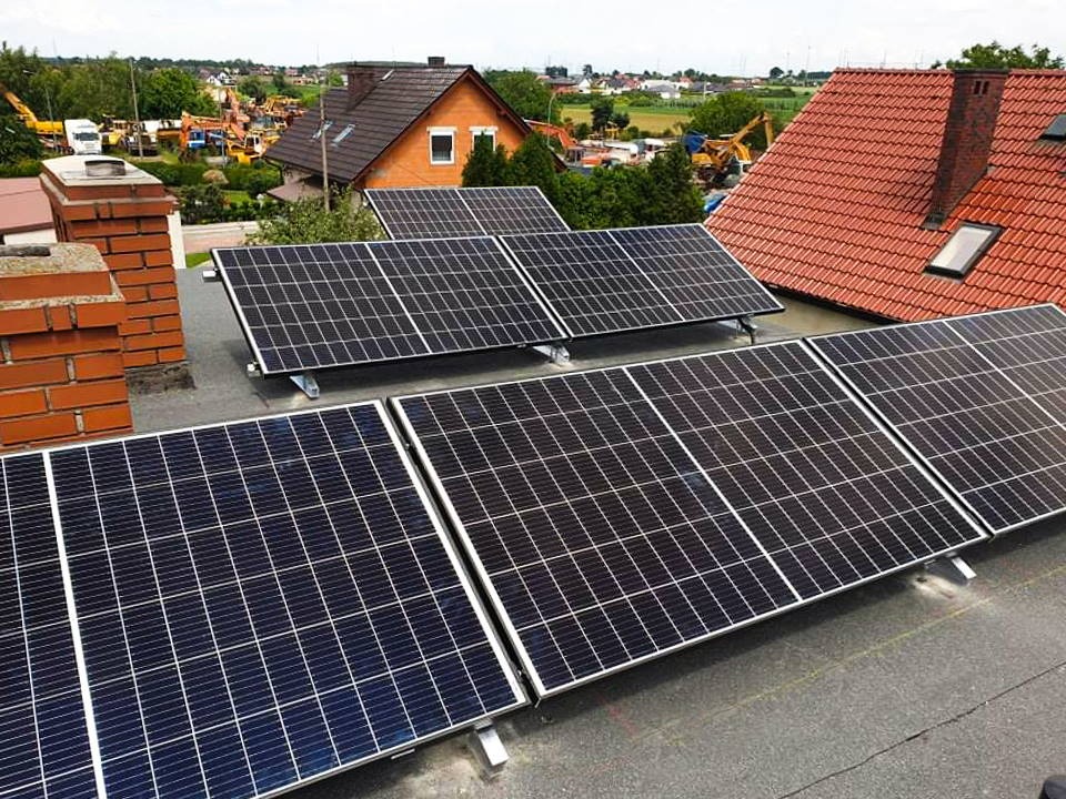 Panele słoneczne zamontowane na płaskim dachu budynku, z widocznymi kominami z cegły i dachami sąsiednich domów w tle.