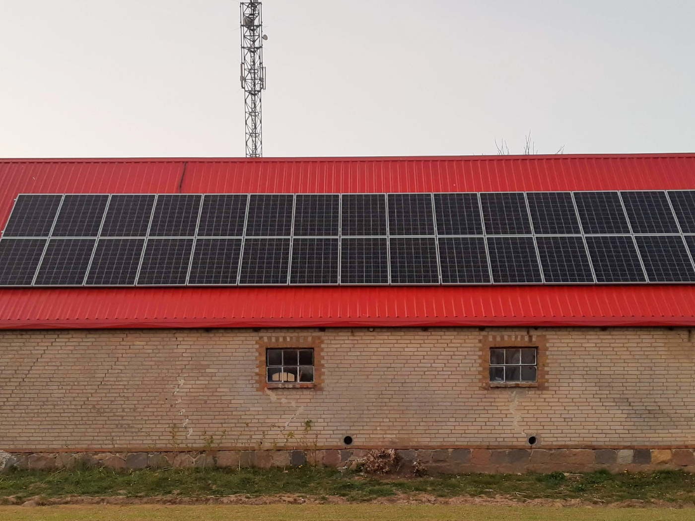 Rząd paneli słonecznych na czerwonym blaszanym dachu budynku z cegły. W tle maszt telekomunikacyjny.