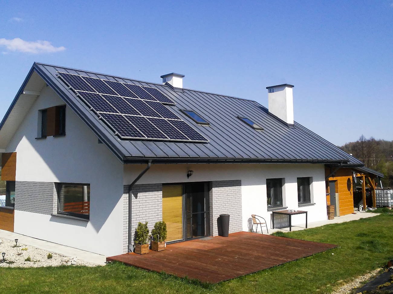 Nowoczesny dom jednorodzinny z panelami słonecznymi na szarym dachu blaszanym, widoczne okna dachowe, biała elewacja z elementami cegły i drewna, taras z drewna, trawnik.