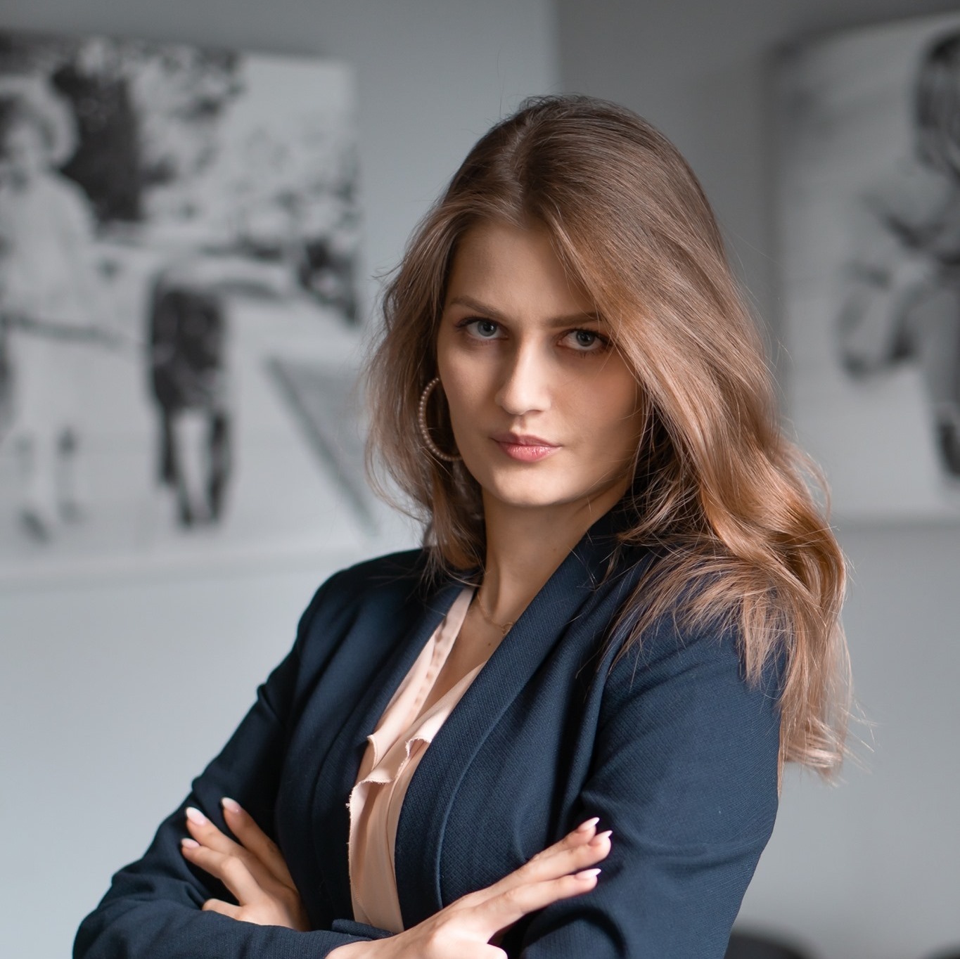 Portret kobiety w granatowej marynarce i bluzce w odcieniu pudrowego różu, z rękami skrzyżowanymi na piersiach, patrzącej wprost, z rozmytym czarno-białym zdjęciem w tle.